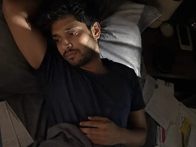 A young man lying awake in bed at night, bills and papers scattered beside him
