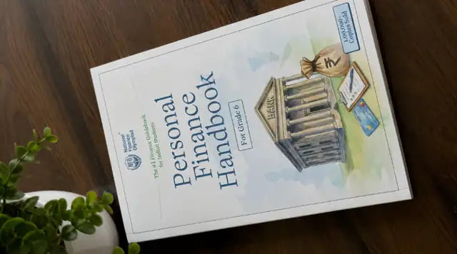 The Personal Finance Handbook for Grade 6, on a wooden desk next to a small plant